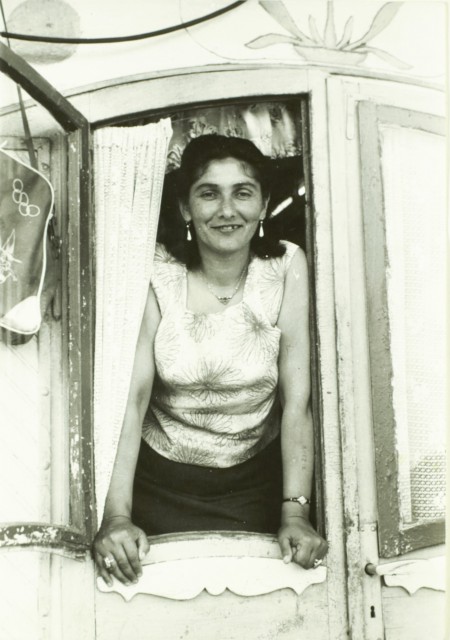 A woman with short dark hear wearing a white top leans out of a window, smiling