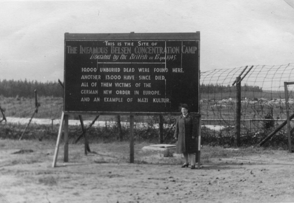 A woman stands in front of a sign erected outside Bergen-Belsen after liberation, 1945